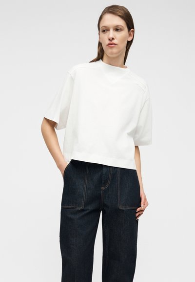 Γυναίκα που φοράει άνετο λευκό κοντομάνικο τοπ και σκούρα μπλε τζιν ψηλόμεσο, στέκεται με το ένα χέρι στην τσέπη, κοιτώντας προς τα αριστερά.