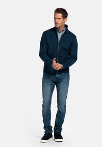 Marineblauwe ritsjack in een gladde stof, gecombineerd met een gestreept shirt, blauwe jeans en zwarte sneakers, staand tegen een witte achtergrond.