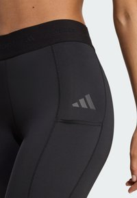 Leggings atletici neri con una fascia in vita ampia e il marchio "TECHFIT" in rilievo. Logo a tre strisce di colore grigio riflettente sul lato.
