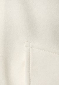 Tissu beige clair à finition texturée, avec une poche ornée de surpiqûres contrastantes sur le côté droit.