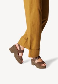 Sandales à plateforme marron avec une semelle texturée et des sangles larges. Associées à un pantalon moutarde avec des accents orange aux poignets.