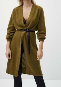 Kvinna i olivgrön lång cardigan med svart bälte över svarta byxor och svart spetsblus, stående mot en enfärgad bakgrund.