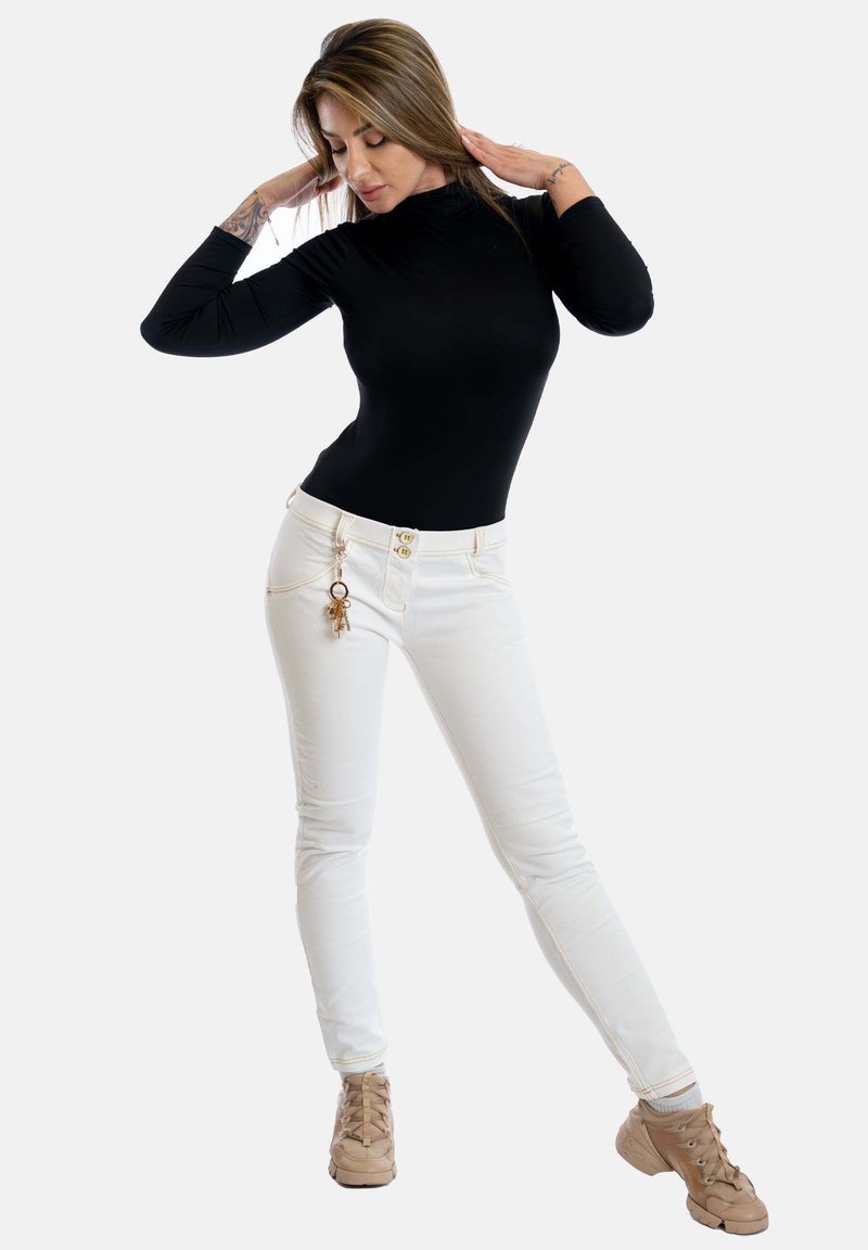 Schwarzes Langarmoberteil, weiße Slim-Fit-Jeans mit goldenen Knöpfen und einem Schlüsselbund-Detail, kombiniert mit beigen Schnürschuhen. Einfache Hintergrund.
