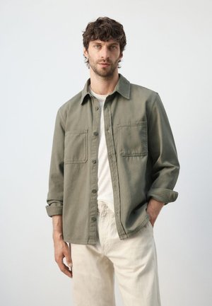 Man with short curly hair wearing an olive green button-up shirt over a white t-shirt and beige pants, standing with one hand in pocket.