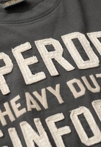 Zwart-grijze zware katoenen t-shirt met witte geborduurde letters die "SUPERIOR HEAVY DUTY" en "SENFORD" zeggen, met een gestructureerde stof.