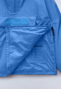 Lyseblå Napapijri-jakke med åben lynlåslomme, der viser inderfor og vaskeanvisningsmærkat med stofegenskaber og brandlogoer.