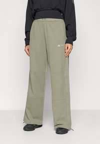 Persona, kas valkā brīvus, olīvzaļus platākā griezuma bikses ar elastīgu jostu un nelielu balto logotipu uz augšstilba, apvienojot ar melniem sneakeriem un melnu topu.