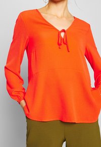 Top lungo arancione brillante con scollatura a V e dettagli con fiocco sul collo. Tessuto testurizzato con polsini arricciati, abbinato a pantaloni verde oliva.
