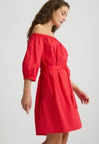 Punane õlatu dress, millel on kolmveerandvarrukad; kerge kangas, vöökoht on kokku tõmmatud nööriga ja voolav seelik.