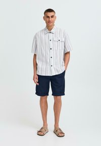 Kurzärmliges, gestreiftes Hemd in hellen Farben mit dunklen Knöpfen, kombiniert mit marineblauen Shorts und hellbraunen Sandalen, ausgestattet mit einer einzelnen Brusttasche.