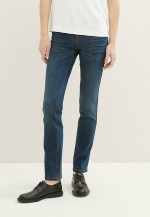 Un jean en denim bleu foncé présente une coupe ajustée avec une taille mi-haute. Associé à des chaussures en cuir noir, mettant en avant une texture lisse et un design minimal.