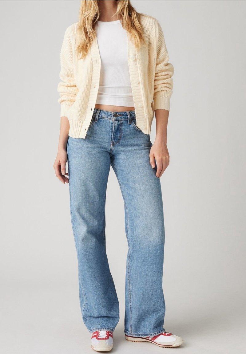Personne portant un cardigan crème, un crop top blanc, un jean large bleu clair et des baskets blanches à rayures rouges, debout de manière décontractée.