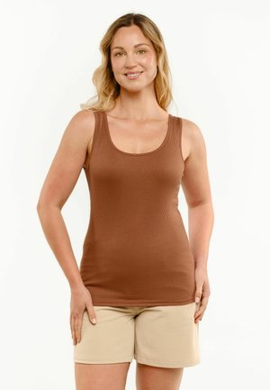 Femme souriante aux longs cheveux blonds portant un haut marron sans manches et un short beige, debout devant un fond clair uni.