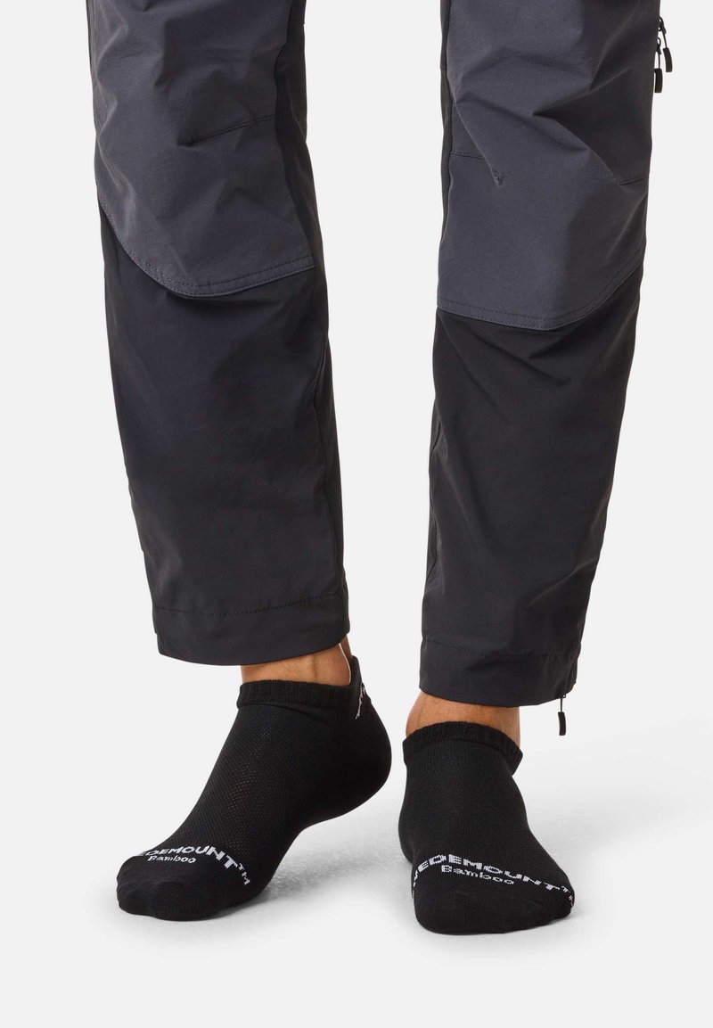 Personne portant des chaussettes noires aux chevilles et un pantalon gris foncé, debout sur une surface claire avec le pantalon légèrement retroussé.