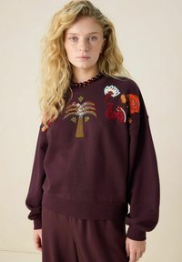 Blonde vrouw die een kastanjebruine trui draagt met kleurrijke geborduurde tropische motieven, staand tegen een effen lichte achtergrond.