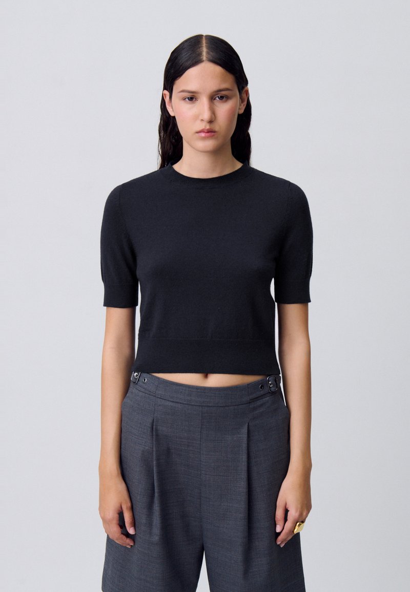 Top de punto negro corto con mangas cortas. Material suave, cuello acanalado y dobladillo. Combinado con pantalones grises con plisados y botones plateados.