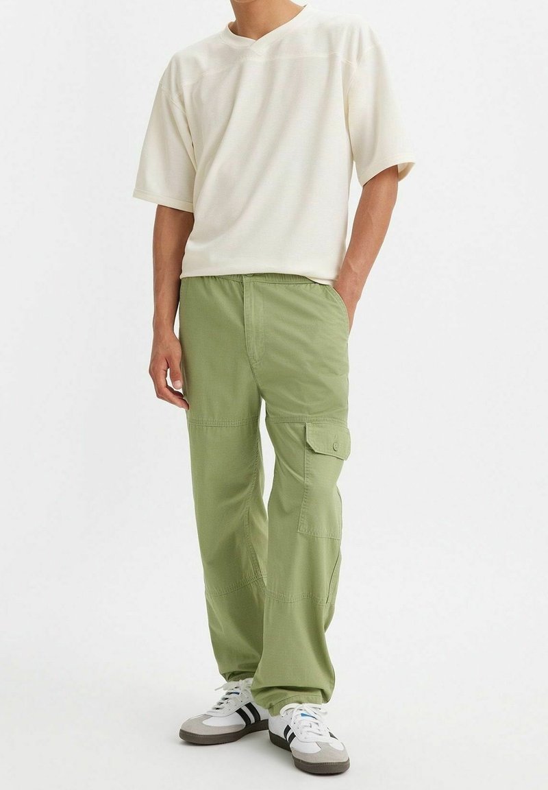 Homme portant un pantalon cargo ample vert clair, une chemise blanche oversize à manches courtes et des baskets blanches avec des rayures noires, debout avec une main dans la poche.
