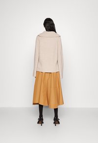 Beiger Strickpullover mit weitem Kragen, kombiniert mit einem midi Rock aus senfgelbem Satin. Das Modell trägt schwarze Strumpfhosen und Riemchenabsätze.