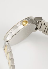 Reloj de metal plateado con un bisel decorado, corona dorada y pulsera de eslabones que presenta secciones alternas de plata y oro.