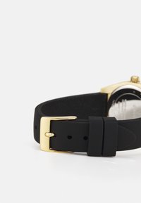 Correa de reloj de silicona negra con hebilla de metal dorado, que presenta una textura suave y una forma redondeada, mostrando parcialmente una esfera de reloj dorada.