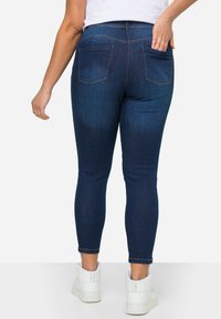 Jeans skinny de mezclilla azul oscuro con una cintura de altura media, que cuentan con dos bolsillos traseros y una textura suave. Combinados con zapatillas altas blancas.
