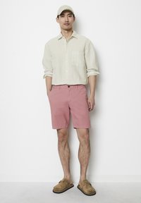 Man wearing a beige cap, cream striped long-sleeve shirt, pink shorts, and brown slip-on shoes standing against a white wall.