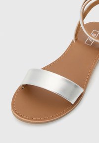 Zilverkleurige metalen sandaal met een bruine faux leren inlegzool. Kenmerkt zich door een brede band over de voorvoet en dunne enkelriemen met stikdetails.
