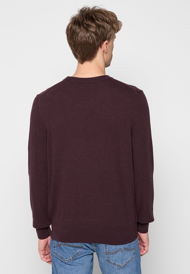 Ein Mann, der sich abwendet, trägt einen dunkelbraunen Pullover und eine blaue Jeans vor einem schlichten hellgrauen Hintergrund.