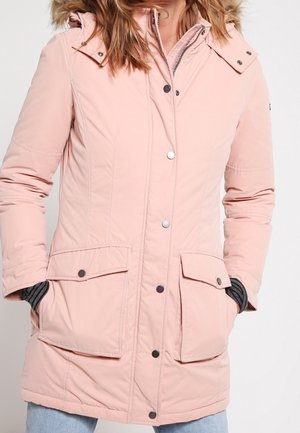 Femme portant un long manteau d'hiver rose clair avec des boutons-pression et de grandes poches avant, les mains partiellement dans les poches, et un pull rayé en dessous.