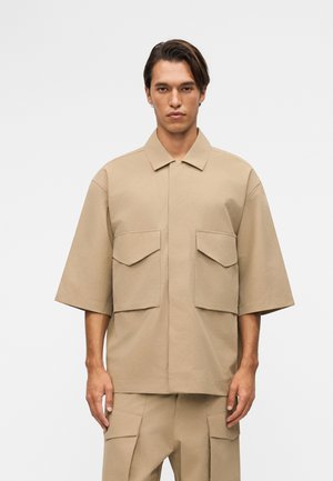 Άνδρας που φοράει μπεζ πουκάμισο με κοντά μανίκια και παντελόνι με μεγάλες τσέπες με καπάκι, στέκεται μπροστά σε λευκό μονόχρωμο φόντο.