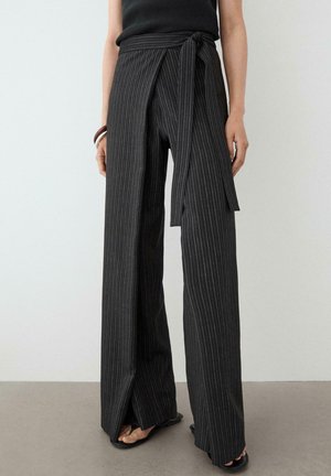 Czarne spodnie w szerokie nogawki w cienkie prążki z wiązanym paskiem w talii, noszone z czarnym topem i sandałami, pokazane od środka tułowia do stóp.