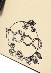 Canvas tas van beige stof, voorzien van een zwart geborduurd logo "Nõbo" en bloemmotieven. Zwarte accenten benadrukken de randen.