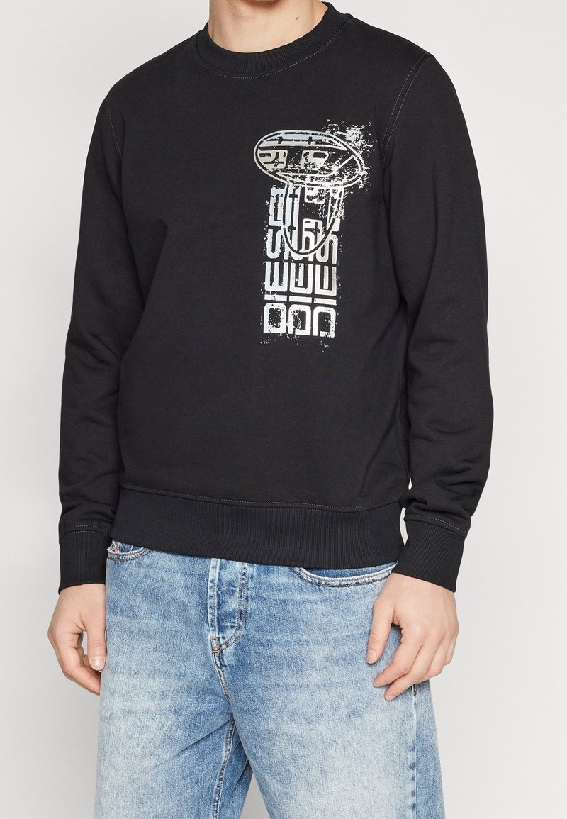 Homme portant un sweat-shirt noir avec un graphique abstrait blanc et un jean bleu délavé sur fond uni.