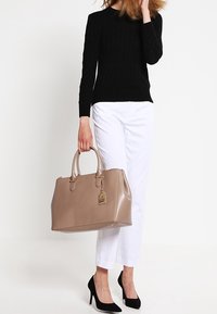 Bolso tote de cuero marrón claro con doble asa, herrajes dorados y una etiqueta decorativa. Combinado con un suéter negro y pantalones cortos blancos.