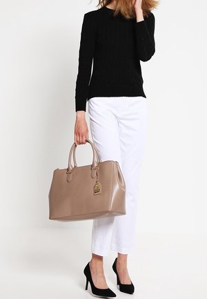 Bolso tote de cuero marrón claro con doble asa, herrajes dorados y una etiqueta decorativa. Combinado con un suéter negro y pantalones cortos blancos.