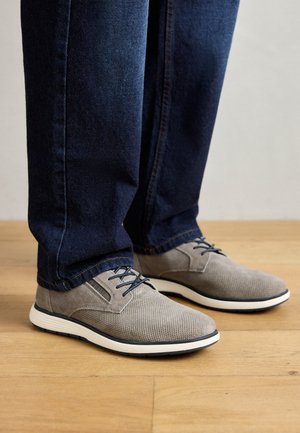 Grau gestaltete Mesh- und Wildlederschuhe mit einer weißen Sohle, die blaue Schnürsenkel und ein strukturiertes Design aufweisen, getragen mit dunklen Jeans auf einem Holzboden.