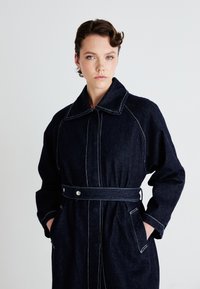 Dunkelblauer Denim-Mantel mit weitem Kragen, weißen Nähten, tailliertem Schnitt und Seitentaschen. Verfügt über eine lockere Passform und lange Ärmel.
