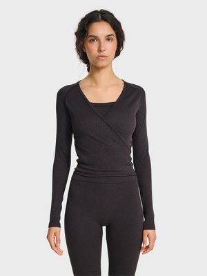 Vrouw met gevlochten haar die een aansluitend donker donker lang mouw overslagtopje draagt en bijpassende donkere legging, staand tegen een effen lichte achtergrond.