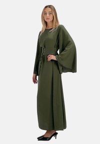 Robe longue vert olive avec des manches larges, taille cintrée et boutons décoratifs. Portée avec des talons noirs. Texture lisse, design longueur de sol.