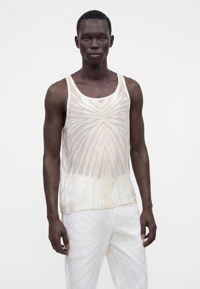 Modèle masculin portant un haut blanc sans manches transparent à motifs et un pantalon blanc assorti à motifs, debout devant un fond blanc uni.