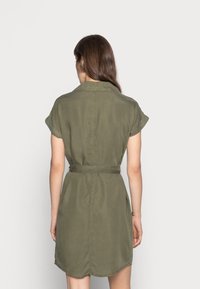 Abito camicia verde oliva, maniche corte, vita con cintura e orlo curvo. Tessuto morbido con una leggera lucentezza e dettagli di cucitura visibili.