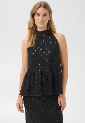 Femme portant un top dos nu noir à sequins et une jupe assortie, debout devant un fond clair uni.