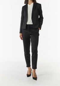 Blazer noir ajusté avec un design structuré, assorti à un pantalon noir cintré et un chemisier blanc. Complété par des chaussures à talons hauts noires.