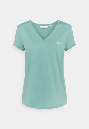 Jasnoturkusowy krótki rękaw z dekoltem w serek, z małą białą haftowaną samolotem na lewej piersi, na jednolitym tle.