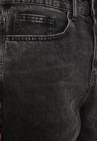 Musta farkut, joissa on kulunut tekstuuri, viisi taskua ja kontrastiväriset ompeleet, jotka näkyvät saumoissa.