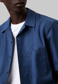 Mies, joka pukeutunut laivastonsiniseen lyhythihainen napilliseen paitaan valkoisen t-paidan päällä, jossa on "ck"-logo rintataskussa.