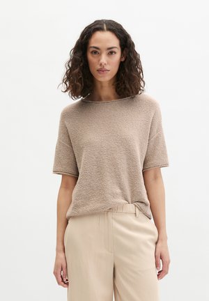 Femme aux cheveux bouclés et foncés, portant un haut beige texturé à manches courtes et un pantalon beige clair, debout devant un fond blanc uni.
