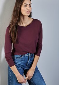 Bordeauxfarbenes Langarmshirt mit einem Rundhalsausschnitt und taillierten Ärmeln, kombiniert mit high-waisted blauen Jeans im klassischen Schnitt.