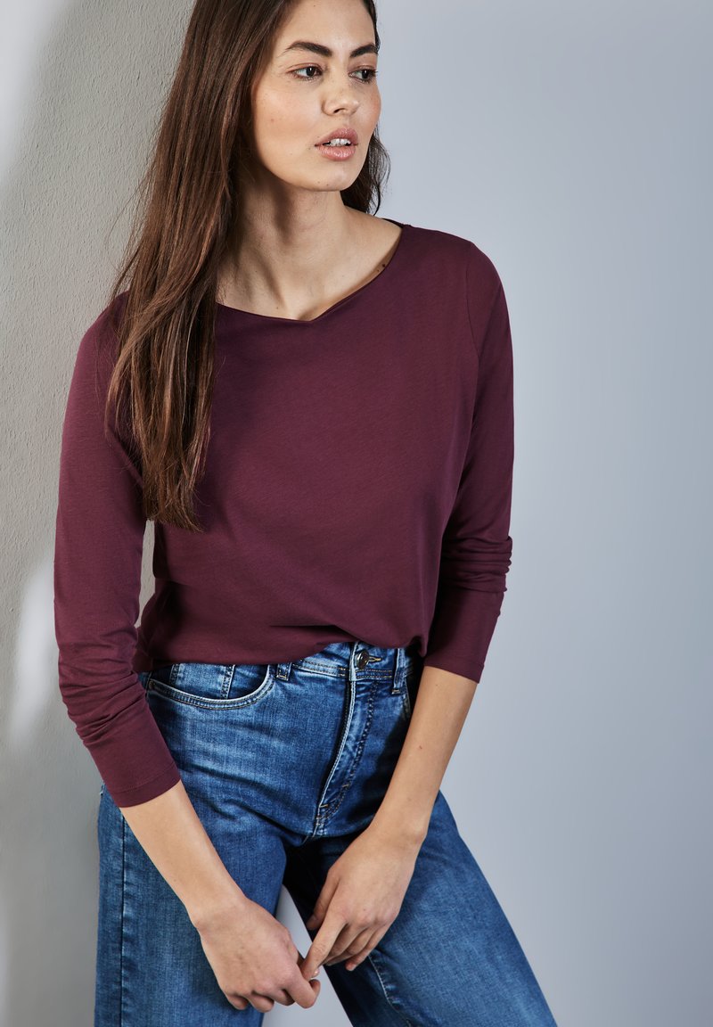 Bordeauxfarbenes Langarmshirt mit einem Rundhalsausschnitt und taillierten Ärmeln, kombiniert mit high-waisted blauen Jeans im klassischen Schnitt.