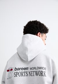 Grå hættejakke med en stor hætte, der har et sort og rødt logo på ryggen, som læser "bareen WORLDWIDE SPORTS NETWORK." Blød stoftekstur.